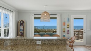 Eine Küche mit einer Granitarbeitsplatte, einer hängenden Korbschwinge, einem Bücherregal und Meerblick durch große Fenster.
