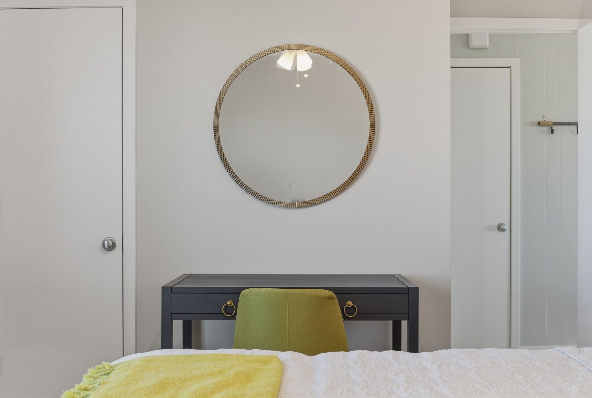 A bedroom with a bed, a bedside table, a green chair, a round mirror, and a door.