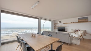 A modern living room with a large window, a dining area, and a sofa. - Apartment Knokke-heist With Sea View (Knokke-Heist)