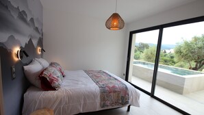 Een slaapkamer met een bed, een houten kruk, een wandlamp en uitzicht op een zwembad door grote ramen.