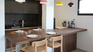 Een moderne keuken met een centraal kookeiland, hangende lampen, en een eethoek met stoelen en een tafel voor twee personen.