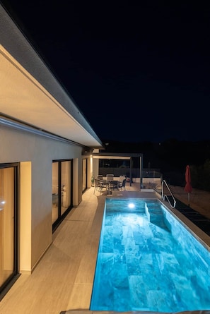 Uma área moderna com piscina ao ar livre, piscina transparente, espaço de refeições com cadeiras e mesa, e um guarda-sol vermelho.