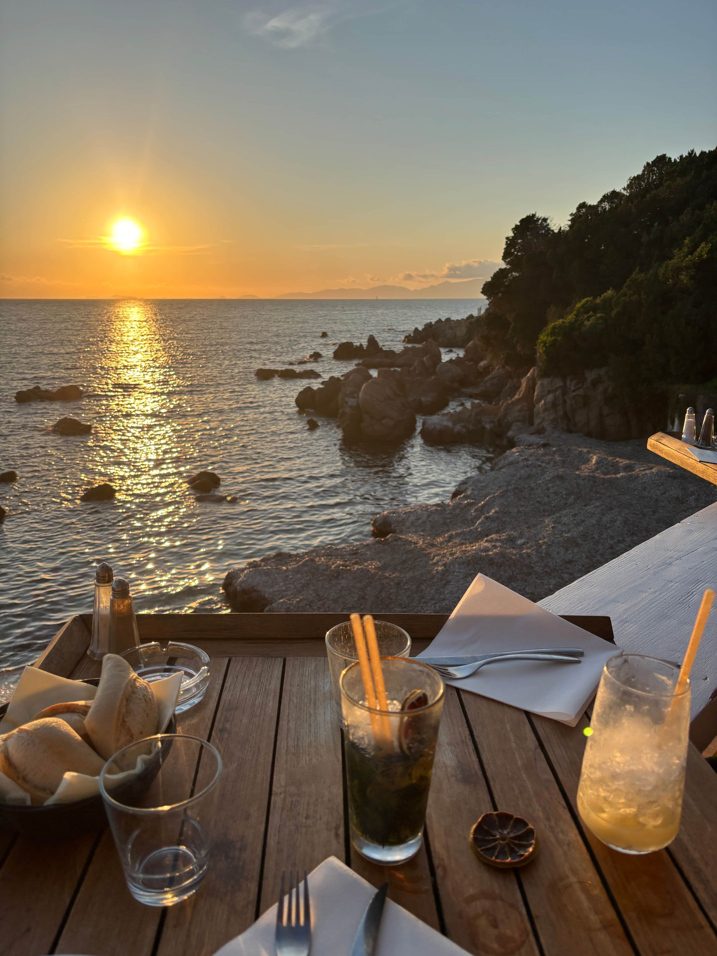 Mesa dispuesta con vasos, una botella y una servilleta, con vista a un litoral rocoso al atardecer.