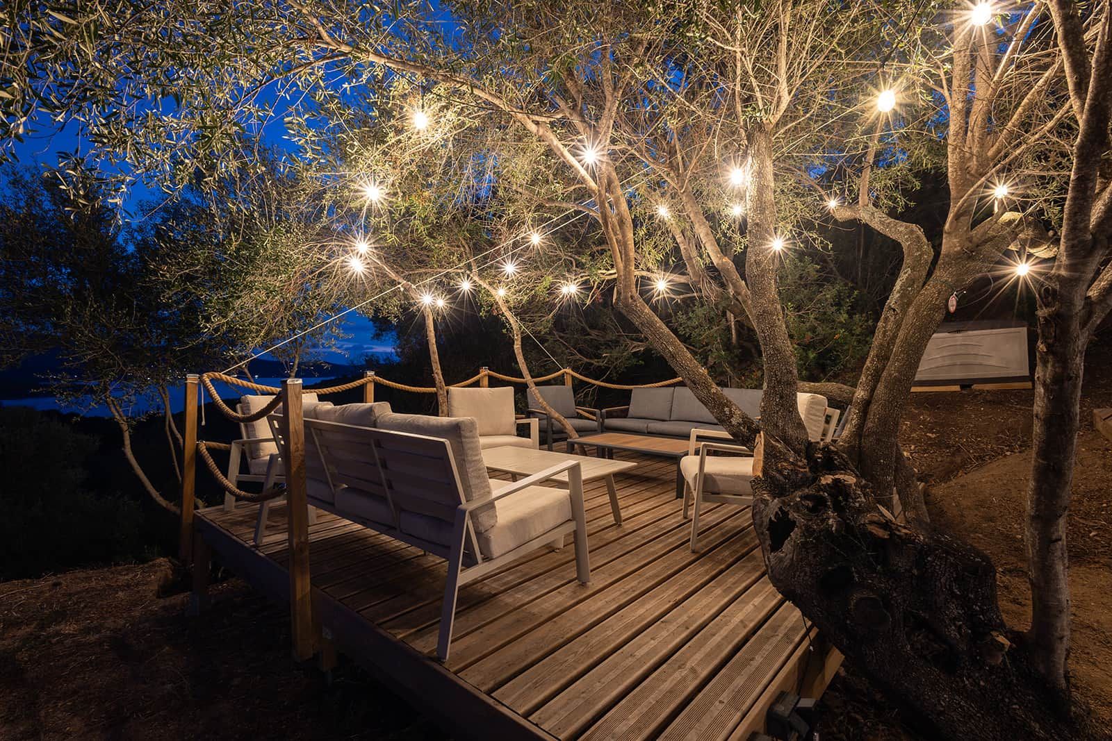 An outdoor seating area with wooden deck and chairs, surrounded by trees and string lights.