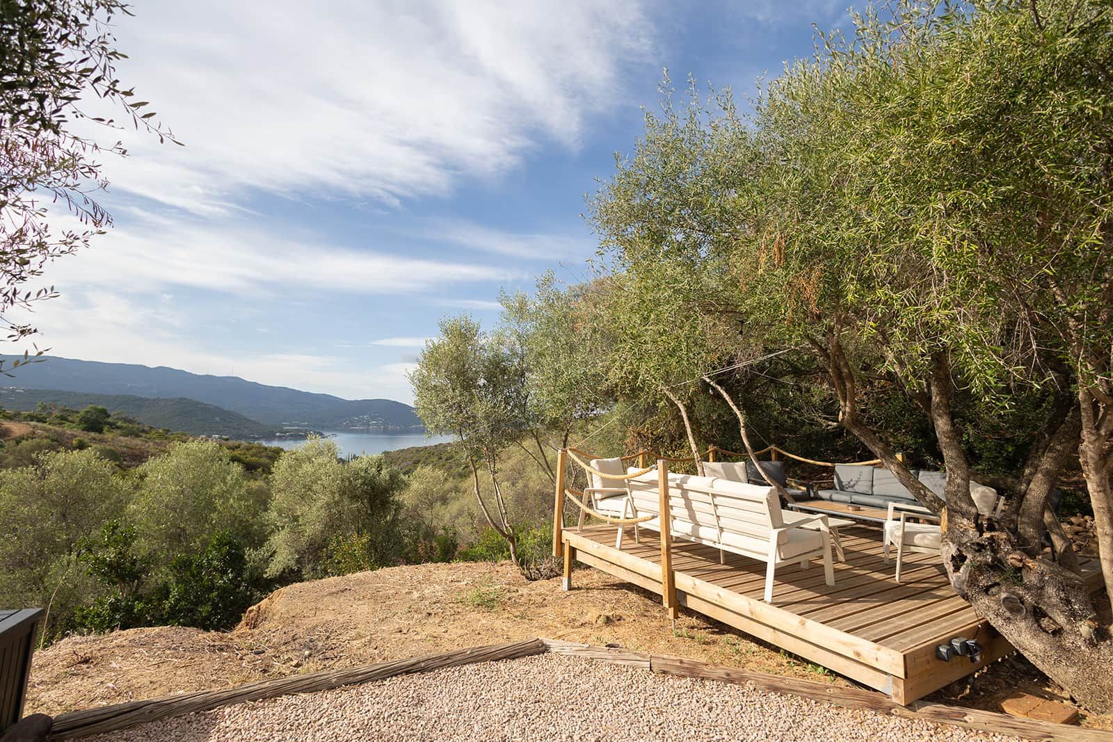 Eine Holzterrasse mit einer Couch und Stühlen mit Blick auf ein Gewässer und Hügel.