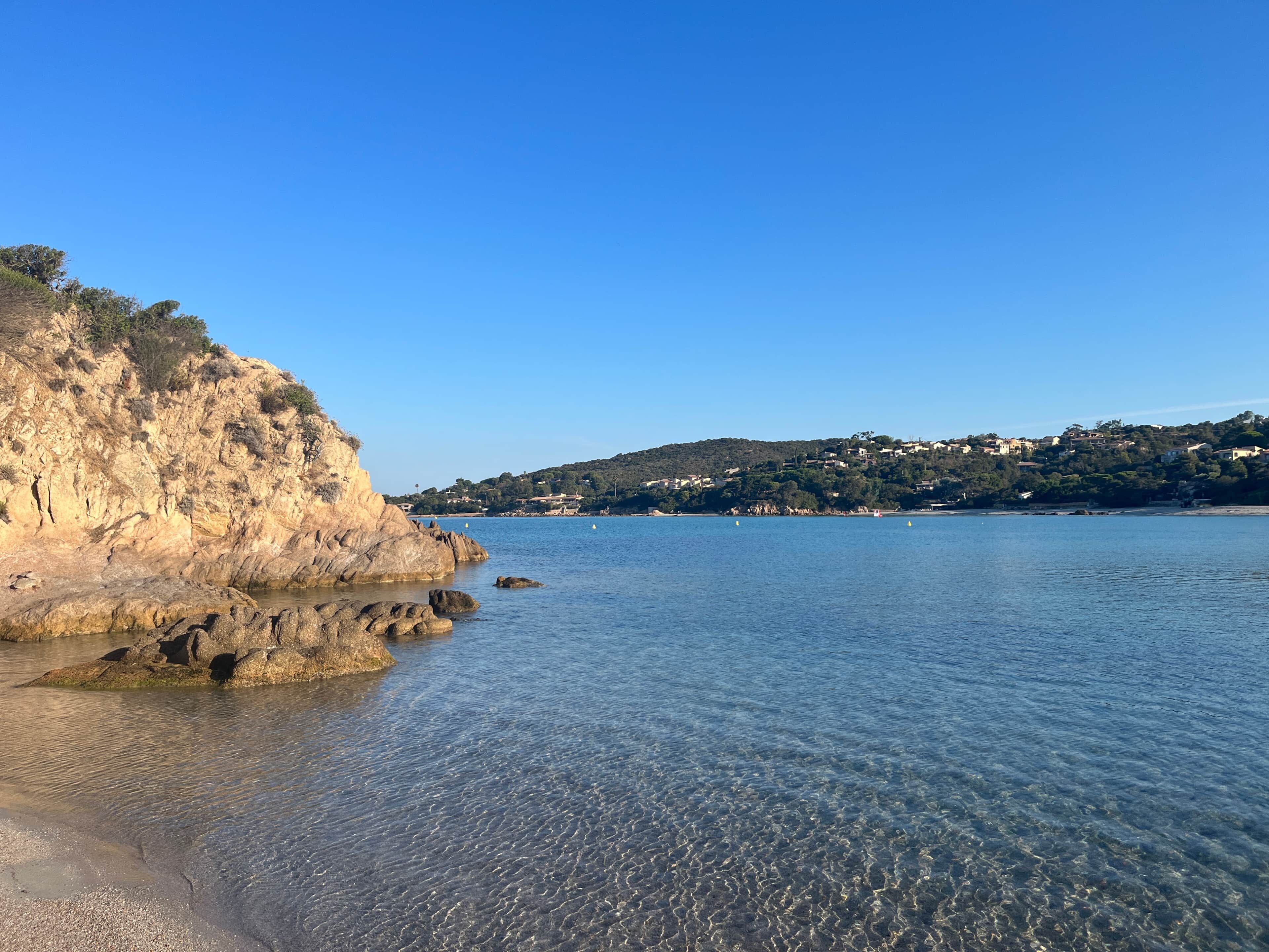A coastal landscape with clear water, rocky cliffs, and a distant hill with buildings.