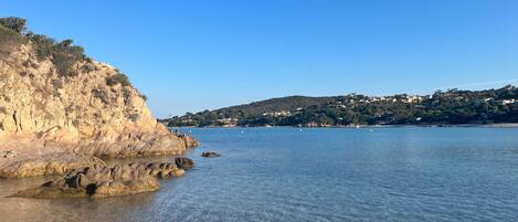 A coastal landscape with clear water, rocky cliffs, and a distant hill with buildings.