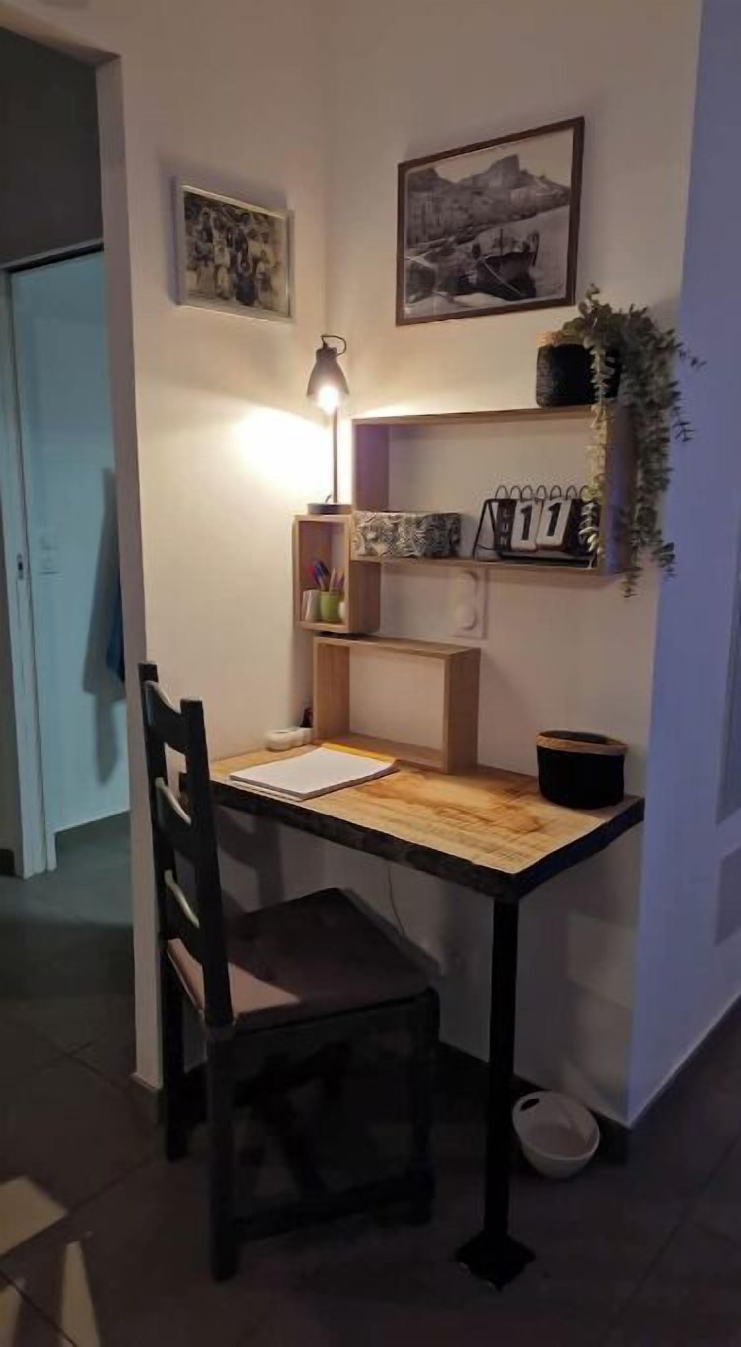 A small, well-lit workspace with a desk, chair, and a lamp. There are shelves with decorative items and a framed picture on the wall.