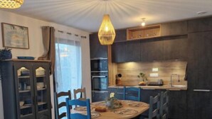 A modern kitchen with dark cabinetry, a central island, and a dining area with a round table and blue chairs. - 70m2 house with large terrace in secure residence with swimming pool. (Bonifacio)