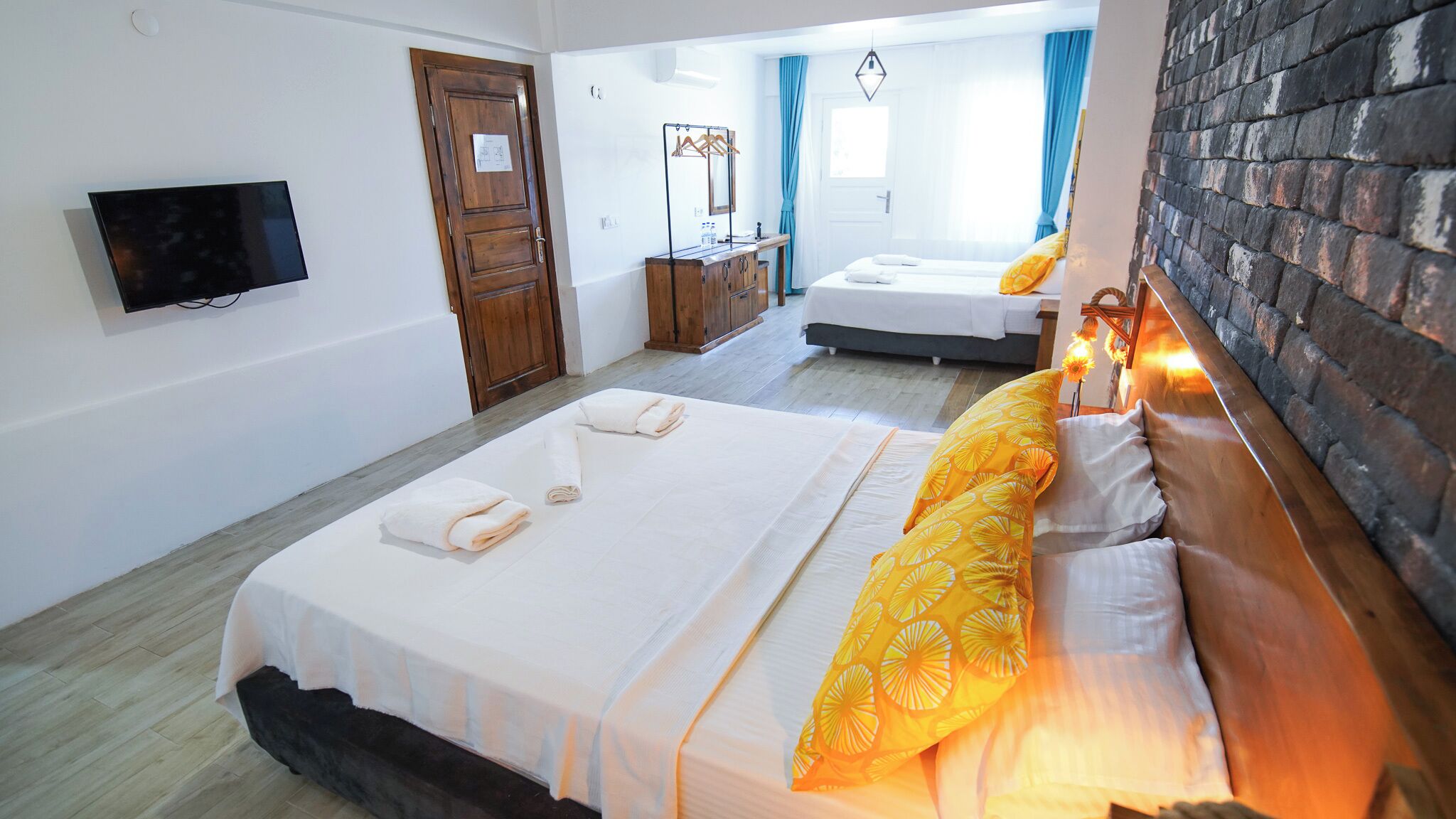 Habitación de hotel con dos camas, un televisor montado en la pared, un armario de madera y una pared de ladrillo.