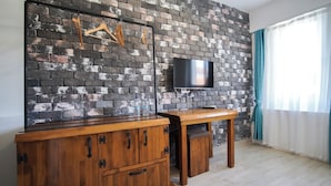 A room with a brick wall, a flat-screen TV mounted on it, a wooden cabinet with drawers, and a window with curtains.