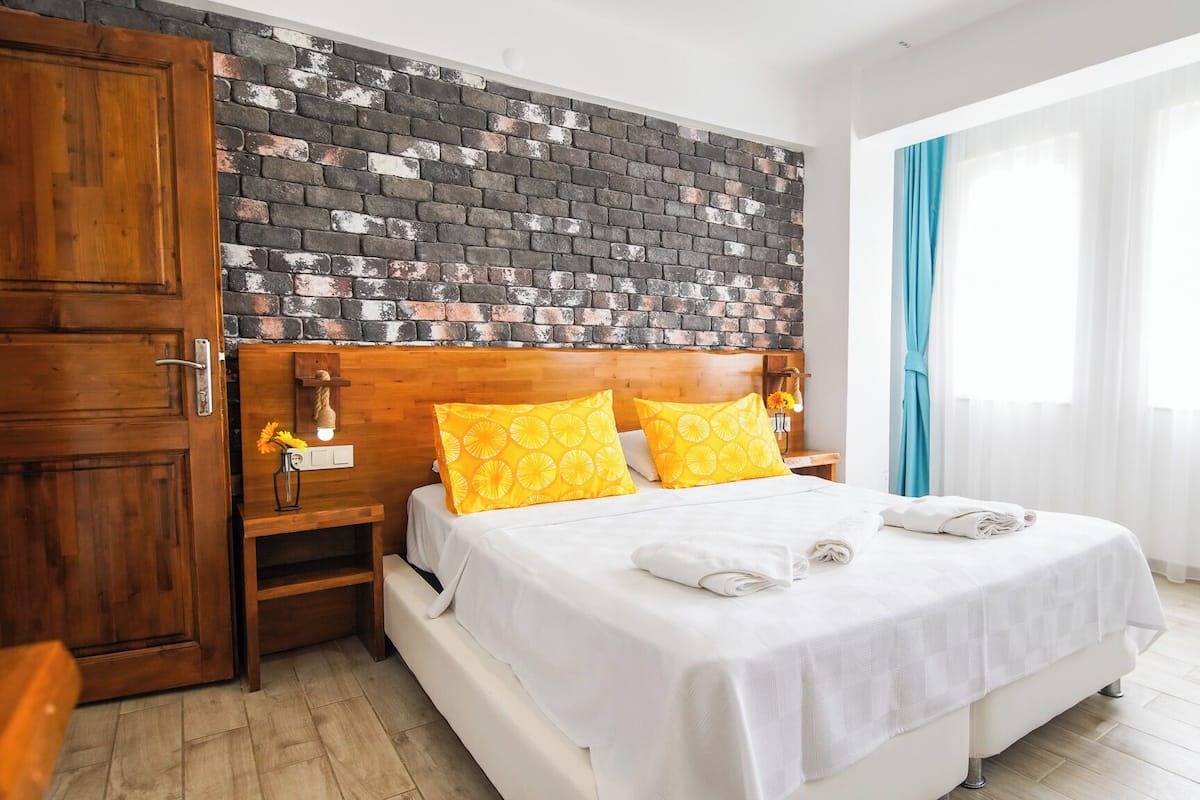 Habitación de hotel con cabecera de ladrillo, puerta de madera, mesita de noche con lámpara, cama con ropa blanca y almohadas amarillas, y ventana con cortinas azules.
