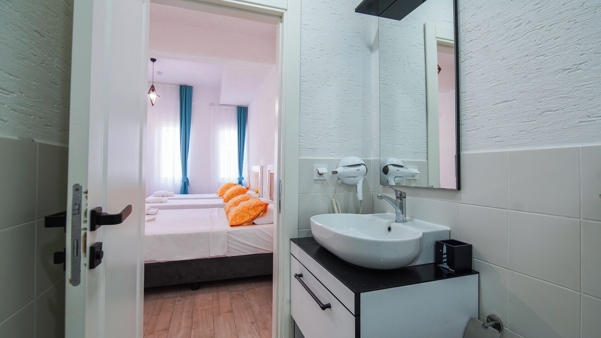 A hotel room with a bed, a bathroom with a sink and mirror, and a view of another room with a bed and curtains.