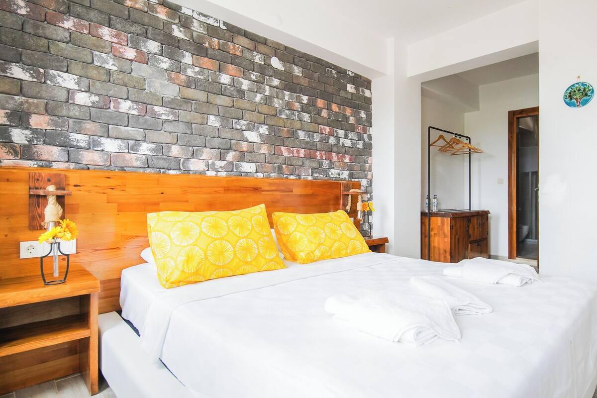 Un dormitorio con cabecera de ladrillo, una mesita de noche de madera con lámpara, y una cama con ropa de cama blanca y almohadas amarillas.