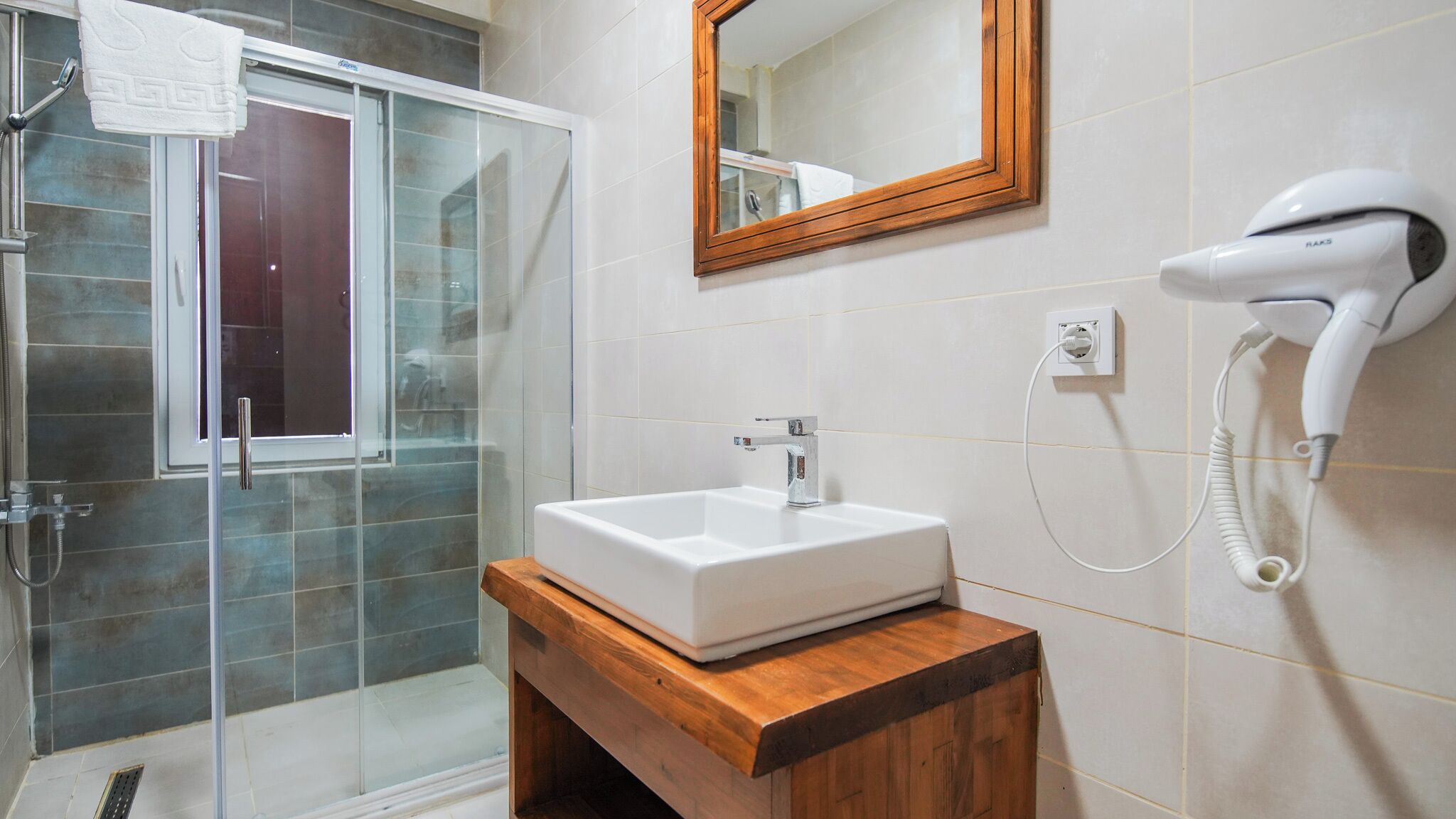 A bathroom with a glass shower enclosure, a white sink on a wooden vanity, and a hairdryer on the wall.