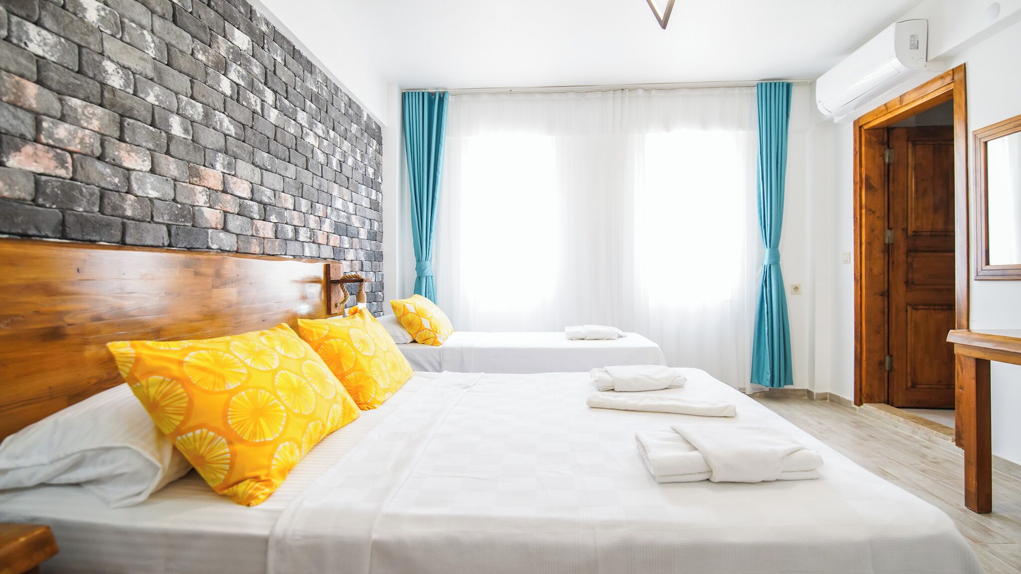 Habitación de hotel con pared de ladrillo, dos camas con sábanas blancas, almohadas amarillas, cabeceros de madera, una puerta de madera y una ventana con cortinas azules.