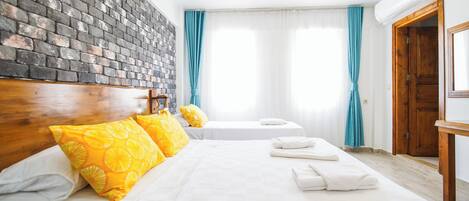 Habitación de hotel con pared de ladrillo, dos camas con sábanas blancas, almohadas amarillas, cabeceros de madera, una puerta de madera y una ventana con cortinas azules.