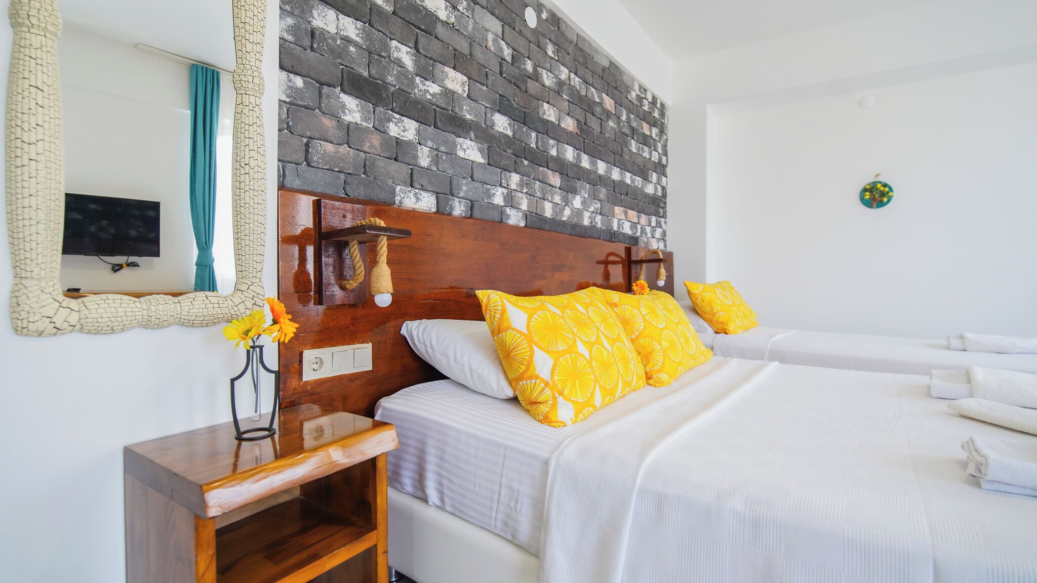 A hotel room with a brick-patterned headboard, a wooden nightstand, two beds with white bedding and yellow pillows, a mirror with a decorative frame, and a small table with a vase of flowers.