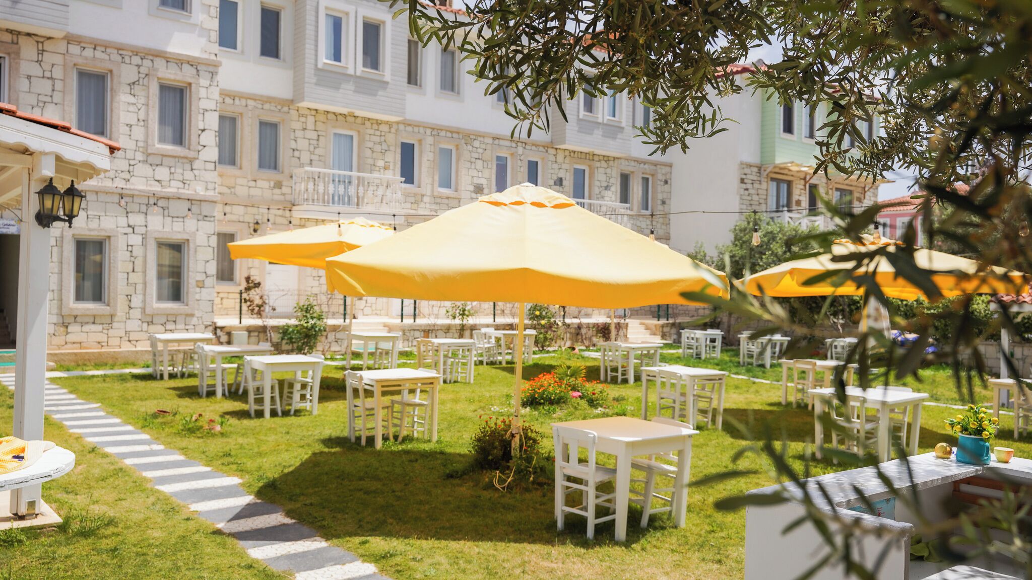 Un espace extérieur aménagé avec des tables et des chaises blanches, des parasols jaunes et un bâtiment en pierre en arrière-plan.