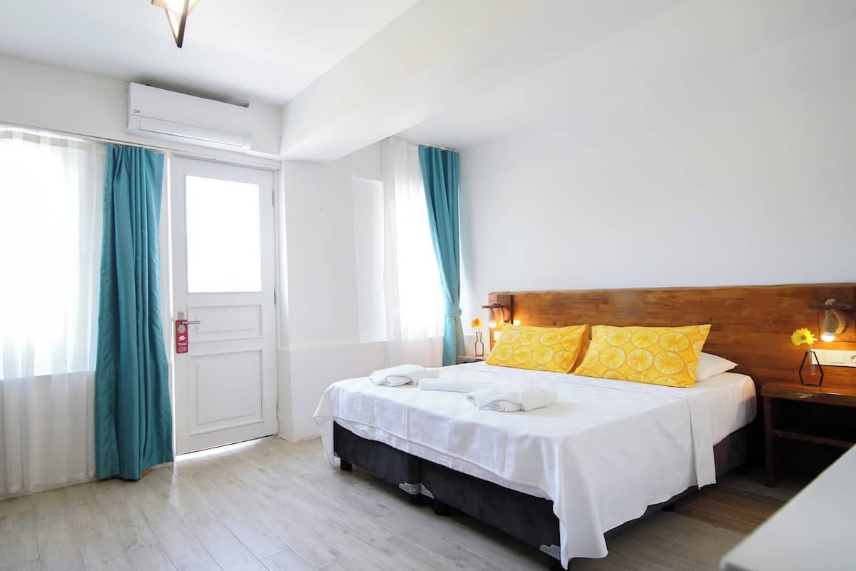 Habitación de hotel con cama, cabecera de madera, mesita de noche, lámpara y ventana con cortinas.