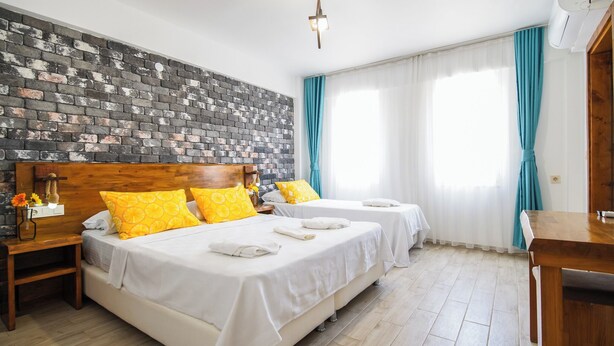 A hotel room with a brick wall, two beds with white linens and yellow pillows, a wooden headboard, a nightstand with a lamp, and a wooden desk.