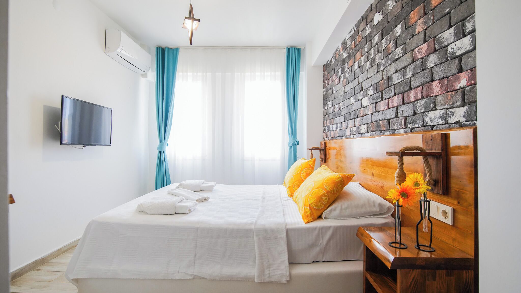 A hotel room with a bed, a TV mounted on the wall, a brick-patterned headboard, and a window with curtains.