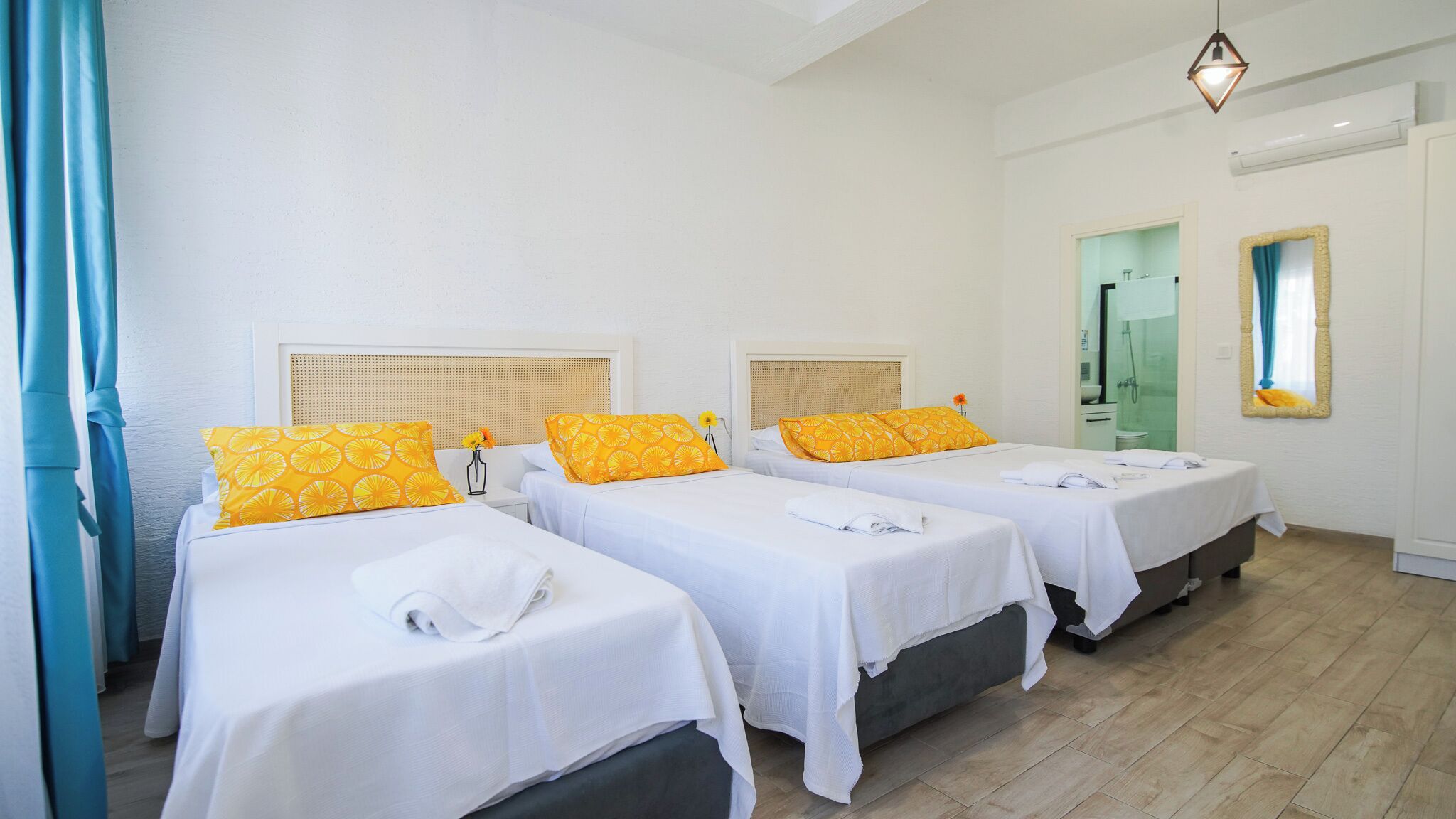 A hotel room with three single beds, each with a white bedspread and a yellow pillow with a circular pattern. There is a small table with a vase of flowers and a lamp. The room has white walls, a wooden floor, and a window with a curtain.