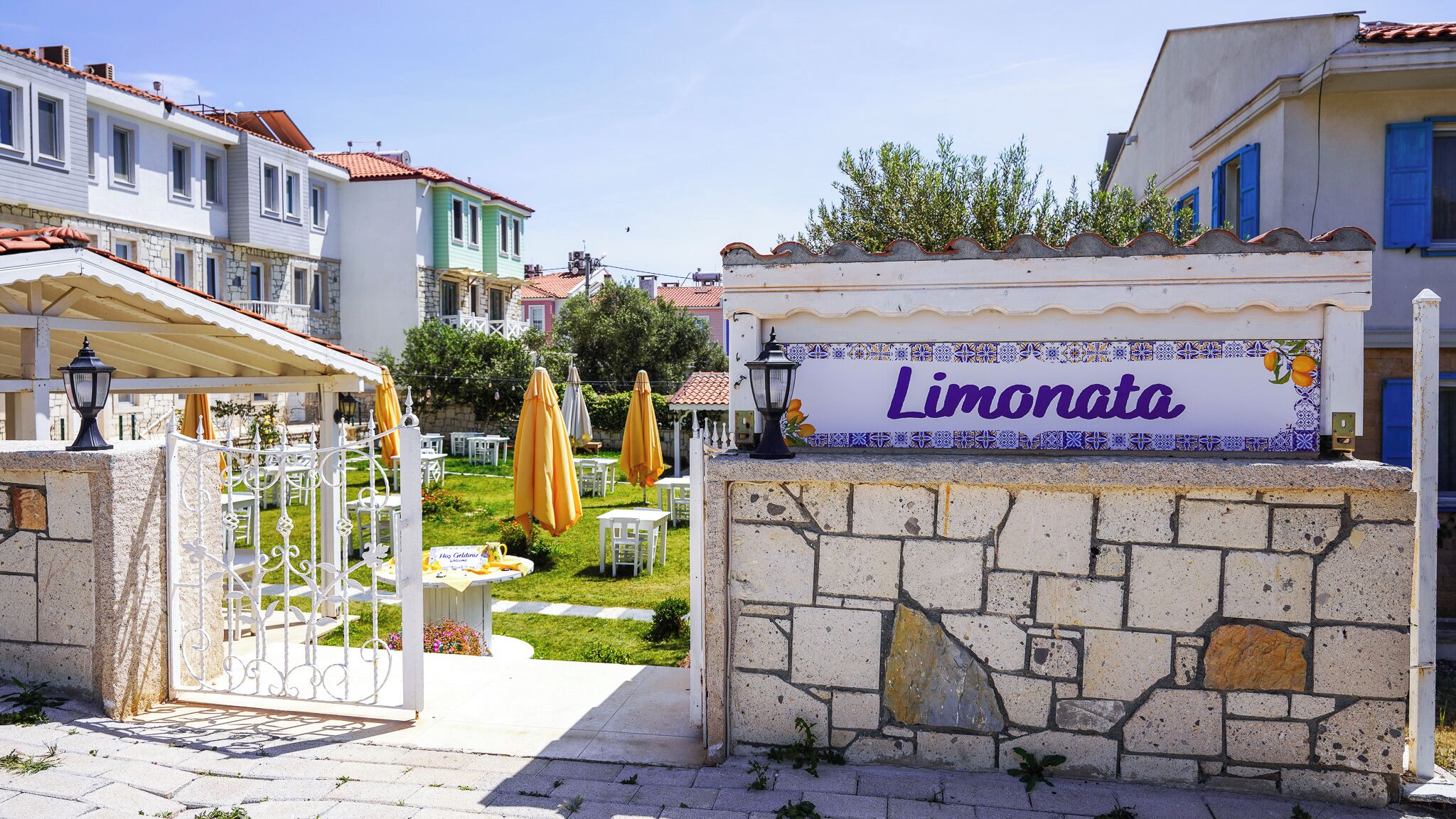 A sign reading "Limonata" at the entrance of a property with a stone wall, a gate, and outdoor seating with umbrellas.
