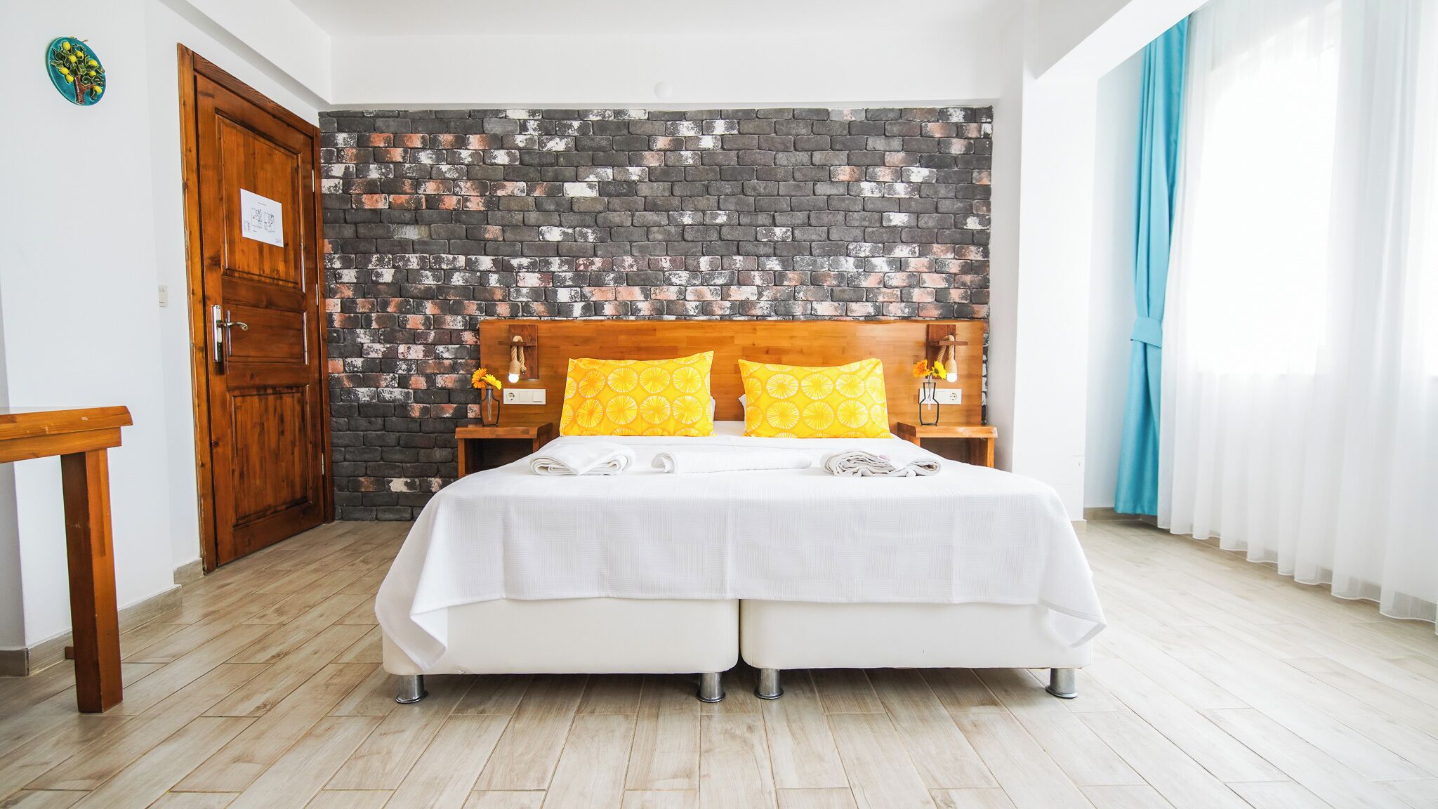 A double bed with white bedding and yellow pillows, a brick-patterned wall, a wooden door, and a wooden nightstand with a lamp.
