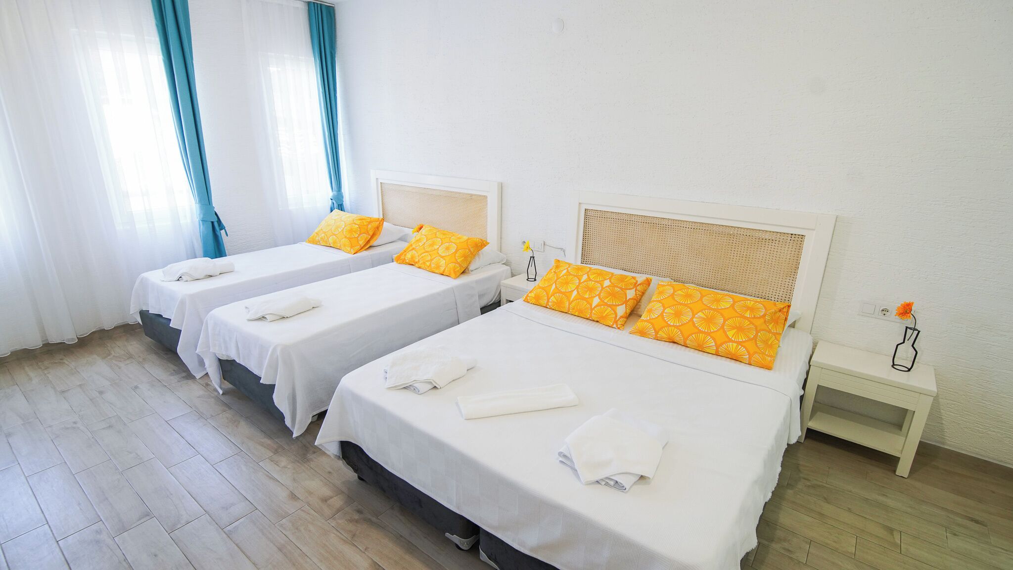 Une chambre d’hôtel avec deux lits simples, chacun agrémenté de draps blancs et d’oreillers à motifs orange. On y trouve une petite table de chevet avec un vase de fleurs et une prise électrique au mur.