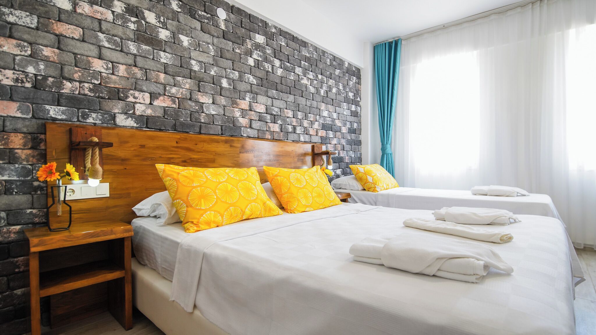 Une chambre d’hôtel avec un mur en briques, deux lits avec du linge de lit blanc, des coussins à motifs jaunes, une table de chevet en bois, un vase avec des fleurs et une fenêtre avec des rideaux.