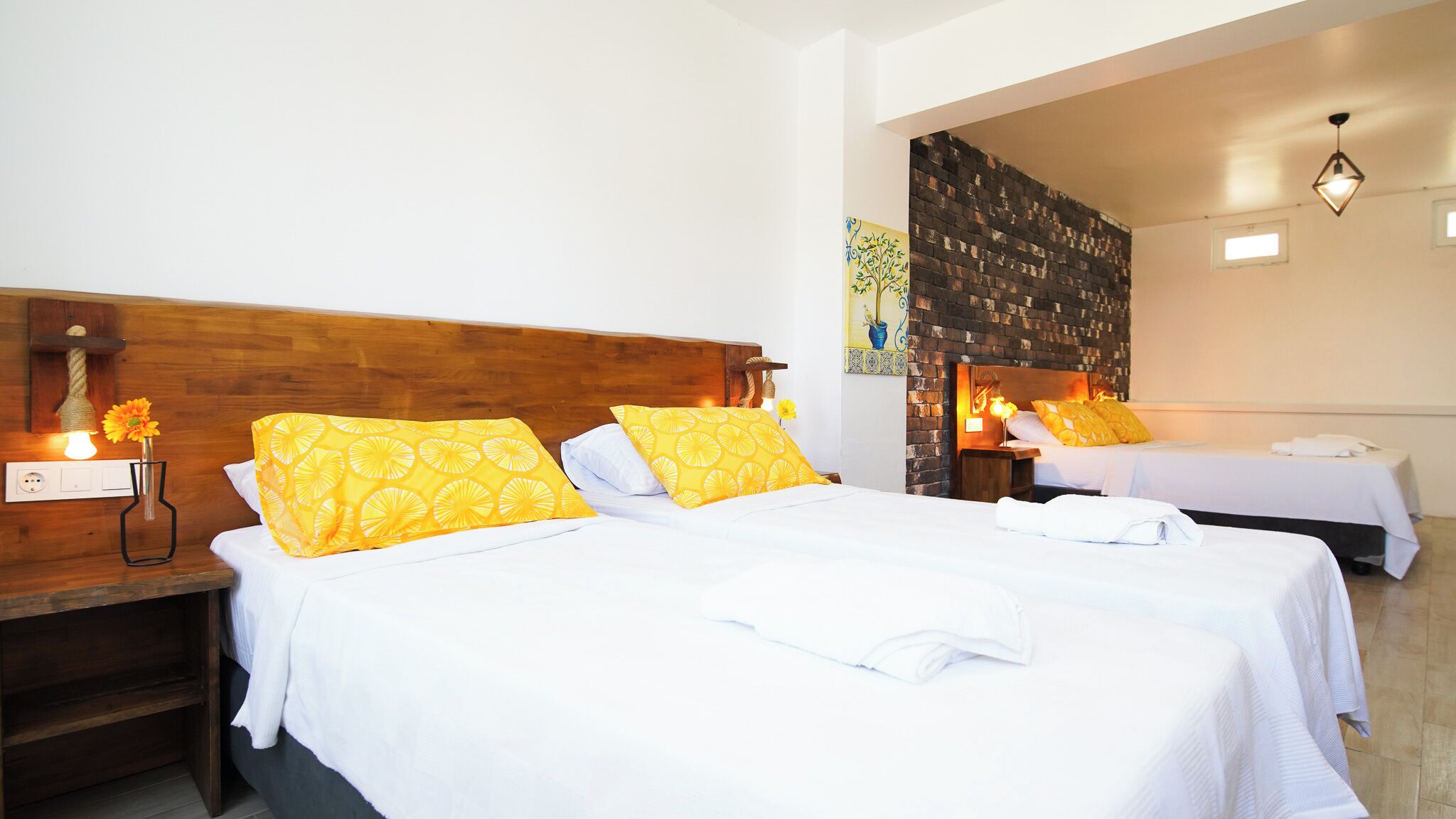 Habitación de hotel con dos camas, cada una con ropa de cama blanca y almohadas estampadas en amarillo. Hay mesitas de noche con lámparas y un aplique de pared.