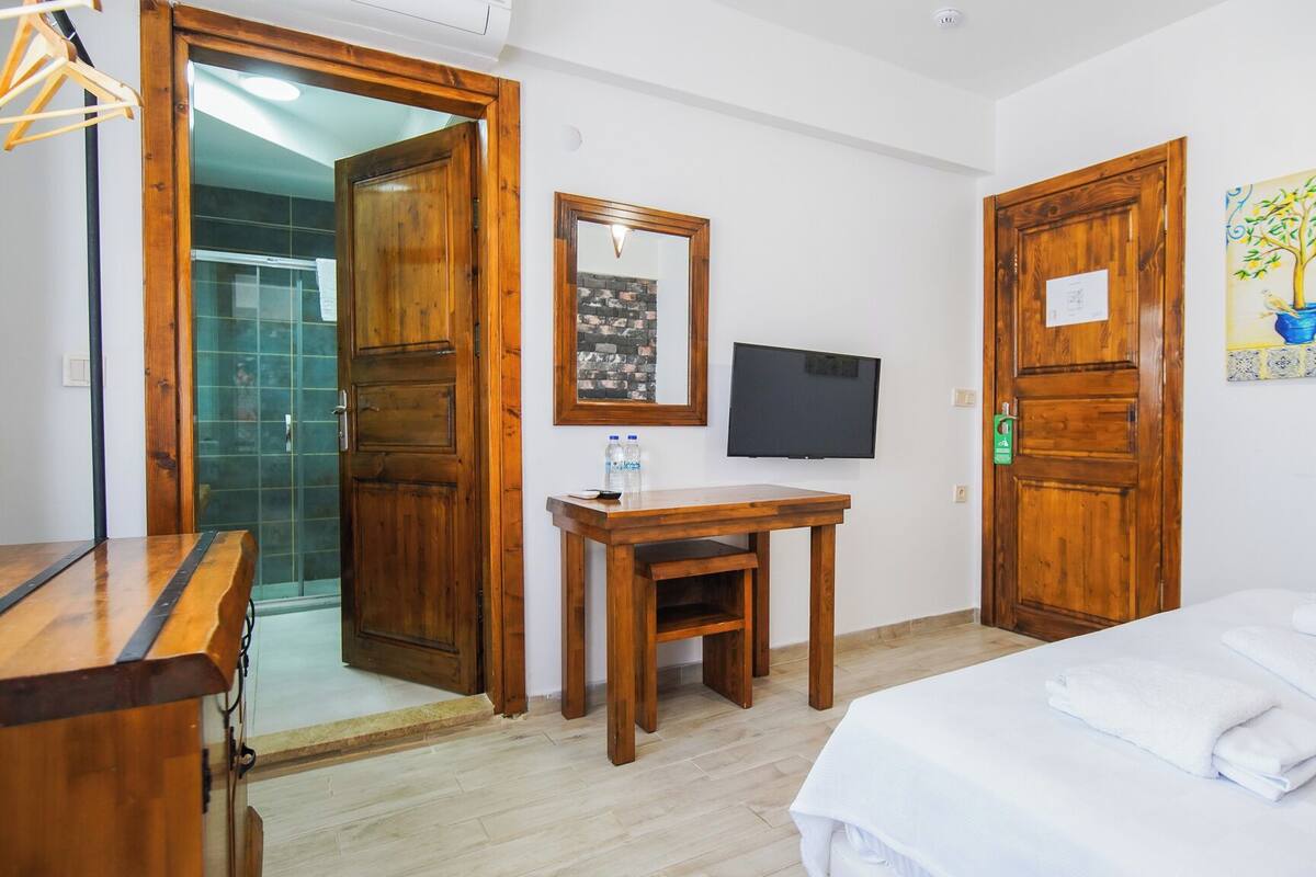 Habitación de hotel con puertas de madera, una cama con ropa blanca, una mesita de madera con una botella, un televisor en la pared y un cuadro de un árbol.