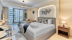 A modern hotel room with a large bed, a seating area with a sofa and armchair, a dining table set for two, and a view of the city through large windows. - bnbme - Gold Crest Views 2 - STUDIO (Dubai)