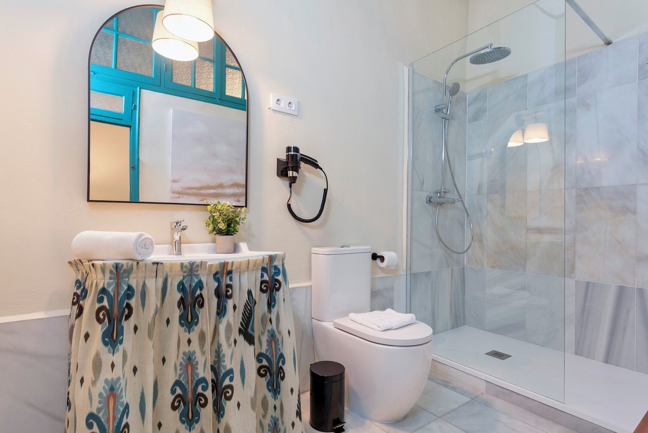 A modern bathroom with a glass shower enclosure, a toilet with a white lid, and a sink with a patterned curtain.