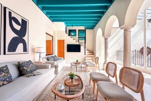 A modern living room with a sofa, coffee table, and chairs, a staircase with a blue ceiling, and a TV mounted on the wall.