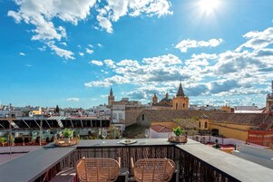 A rooftop terrace with wicker chairs and a table, overlooking a cityscape with historic buildings and a clear sky.