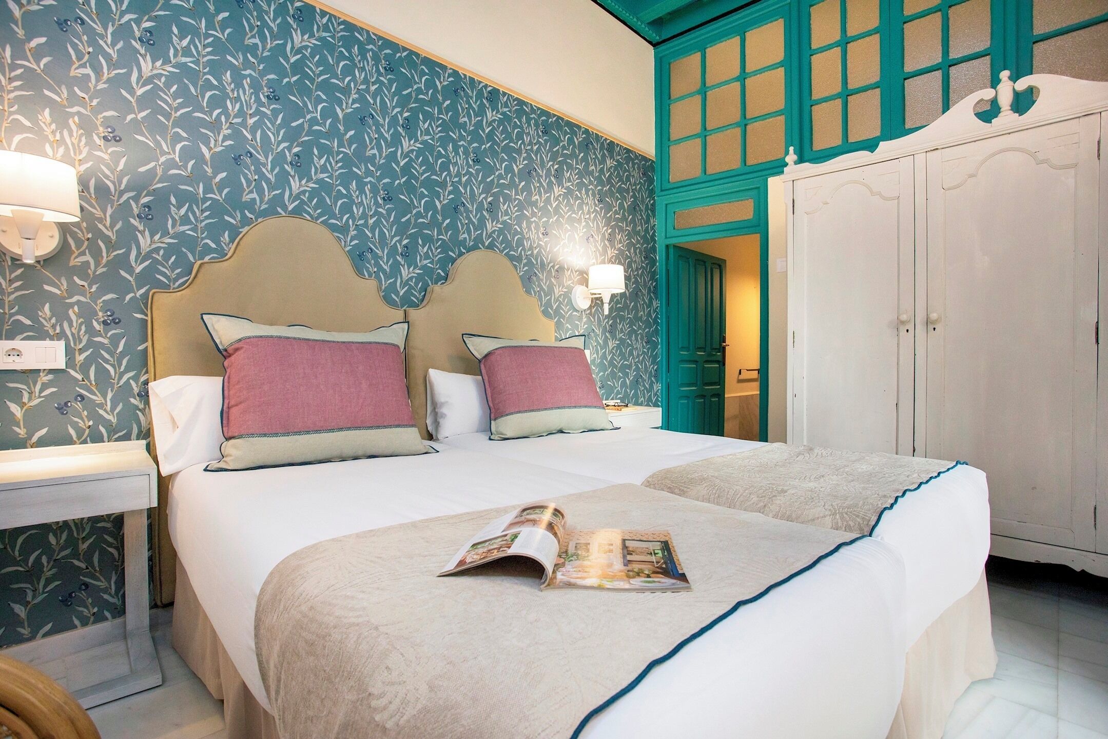 Un lit double bien fait, avec un magazine posé dessus, une table de chevet avec une lampe et une armoire dont la porte est de couleur turquoise.