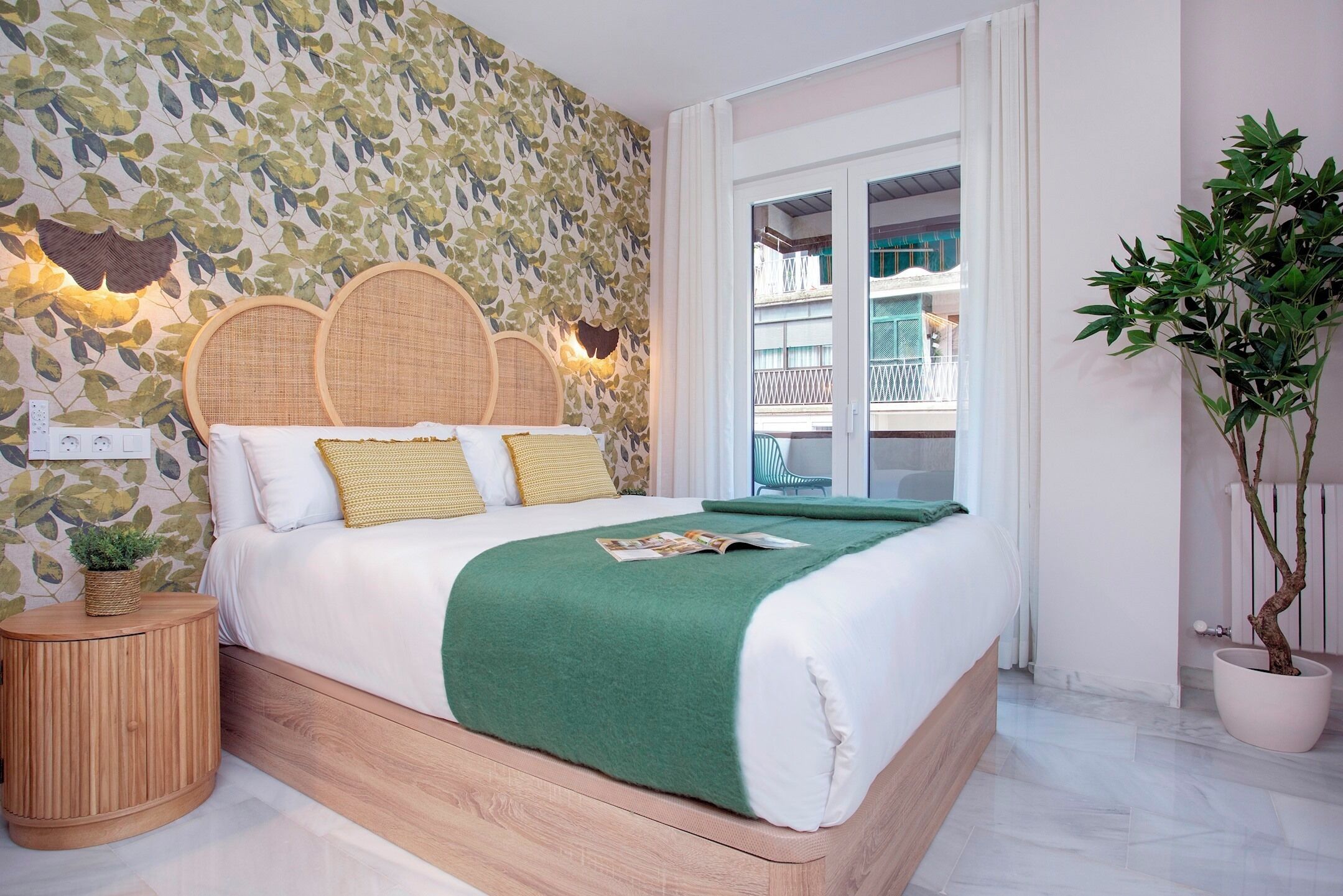 A neatly made bed with a green throw, a wooden bedside table with a plant, and a wall with a leafy pattern.