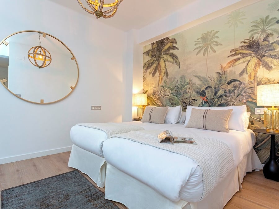 Un dormitorio con una cama grande, un espejo redondo, un mural tropical y dos lámparas de noche.