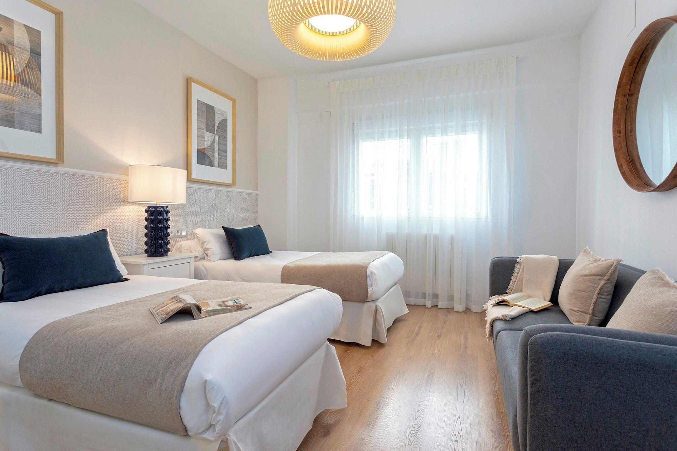 A hotel room with two beds, a sofa, a nightstand with a lamp, and a large window with sheer curtains.
