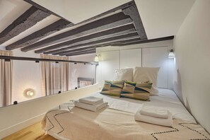A bedroom with a sloped ceiling, a bed with white bedding and patterned pillows, a bedside table, and a window with curtains.