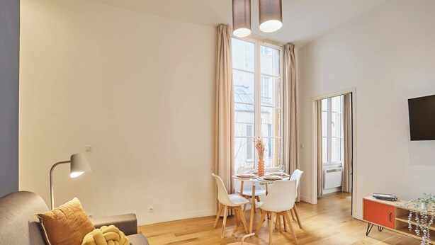 A modern living room with a sofa, a coffee table, a dining table with chairs, a TV mounted on the wall, and a window with curtains. - Superb Apartement Center of Paris Le Marais (Paris)