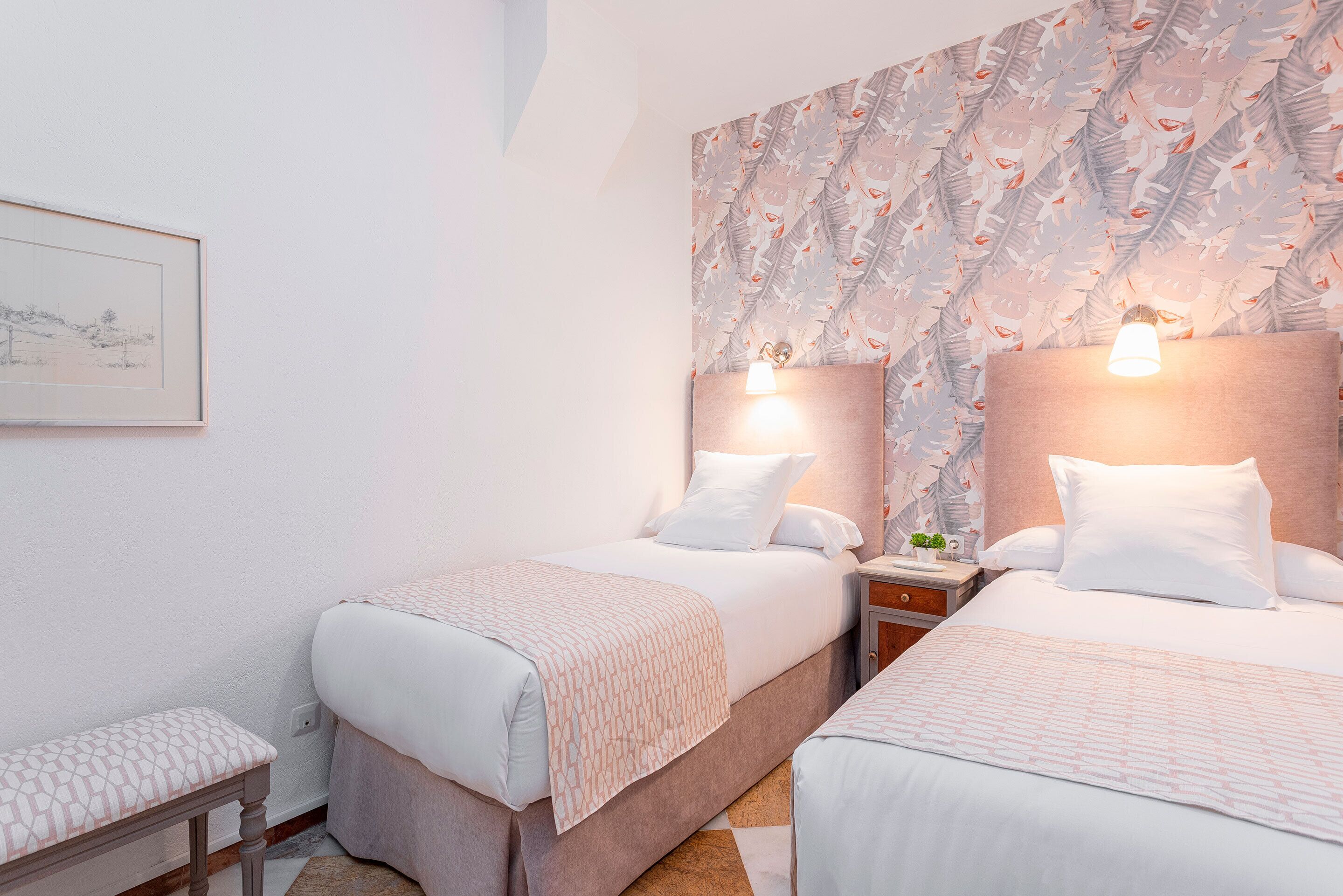 Habitación de hotel con dos camas, un banco, una mesita de noche con lámpara y un cuadro colgado en la pared.