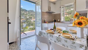 Een eetgedeelte met een tafel gedekt voor twee personen, een keuken op de achtergrond en een balkon met uitzicht op het omliggende landschap.