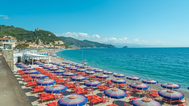 Een strand met rijen rode en blauwe parasols en stoelen, een heldere blauwe zee en gebouwen op de achtergrond.