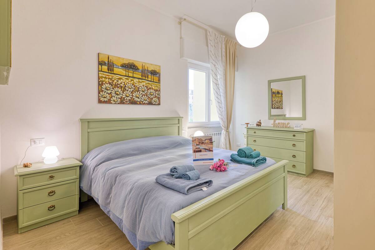 A bedroom with a green bed, a nightstand with a lamp, a dresser, and a window with curtains.