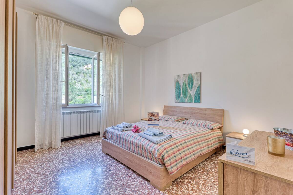 A bedroom with a bed, a bedside table, a nightstand, a window with curtains, a radiator, and a welcome sign.