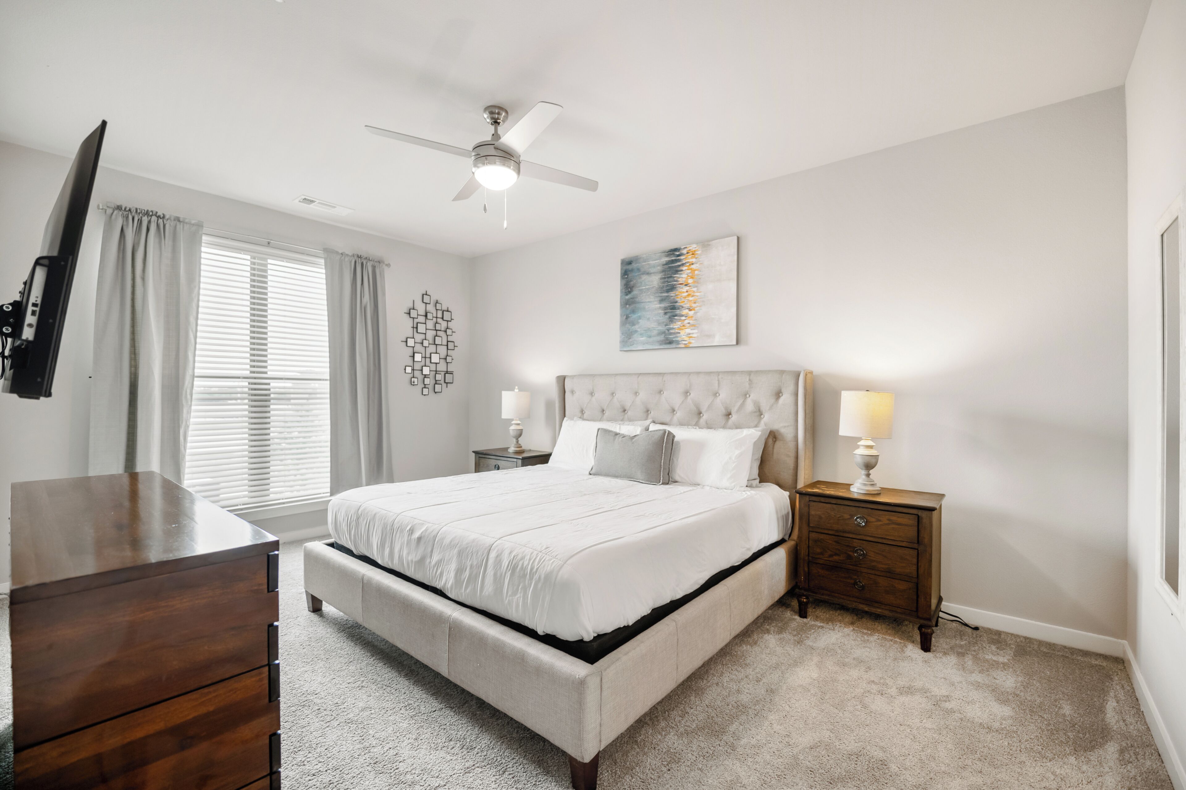 A bedroom with a large bed, a ceiling fan, a TV, a dresser, bedside lamps, and a window with blinds.
