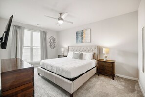 A bedroom with a large bed, a ceiling fan, a TV, a dresser, bedside lamps, and a window with blinds.